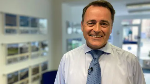BBC News A middle aged man wearing a blue shirt and tie looks towards the camera. He is standing in an estate agent's office