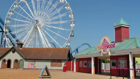 Martin Giles/BBC Buildings are arranged around a courtyard at a theme park entrance. There is a large ferris wheel in the background.