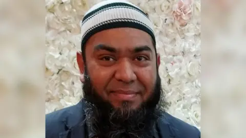 A photograph of Shafiqul Haidhar, a south Asian man, with a long beard and wearing a prayer cap and standing in front of decorate display of white flowers. He is smiling subtly and wearing a smart jacket.
