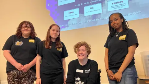 Jon Wright/BBC Four young people in black T-shirts stand with an overhead projection on a wall behind them.