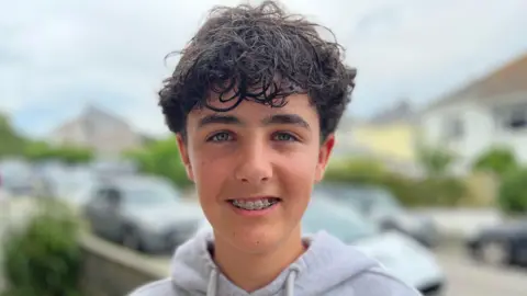 Jacob has short dark tousled hair, he's smiling into camera showing his fixed brace. He has blue yes and is wearing a grey hoodie. There houses and a street in the background.