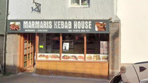 Google The outside of Marmaris Kebab House with the name written on a grey sign above the brown door and window frames