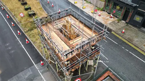 An aerial view of the scaffolding around the statue of King William III on his golden horse. It is surrounded by a platform of wooden planks. There are traffic cones along the road on both sides.
