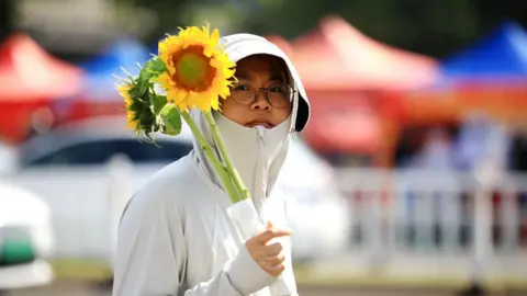 A woman in a thin white jacket zipped up to her chin, holding a bouquet of sunflowers.