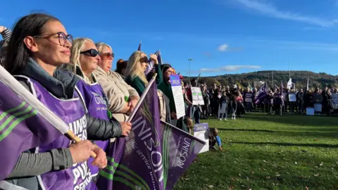 Striking workers - wearing purple Unison tabards - listen to a speech at a rally in a park 