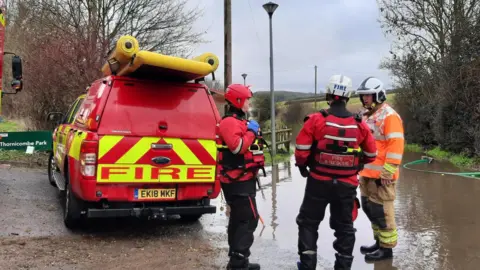 Three firefighters are stood in conversation next to a patch of land flooded with water and a red van with the word 'Fire' printed on the back in large letters. The surroundings are rural, with several bare trees and green fields in the background. To the left of the van is a green sign that reads "Thornicombe Park". 