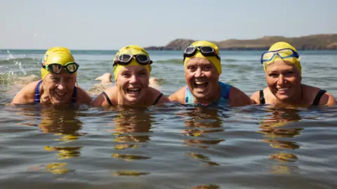 Braving the Blue - Our Lives, BBC Cymru Wales Four women pictured smiling in the sea wearing yellow swim caps and goggles on their head. Their legs are floating on the water's surface. 