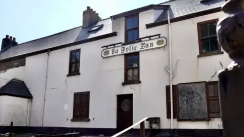 The picture shows the front of an old, abandoned building with a sign that reads “La Folie Inn”. The structure appears to be in poor condition: the windows are boarded up, one of the boards has graffiti on it, and there are visible cracks on the walls. The paint looks faded and chipped, and some debris is scattered near the base of the building. The sign indicates the inn was established in 1722, suggesting it is a historic site.