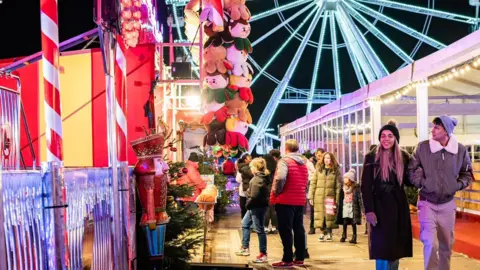 A couple walking past fairground stalls at night, with a ferris wheel in the background. There are people walking in the background.