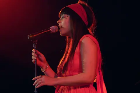 Getty Images Lily Allen wearing a red dress and head scarf singing into a microphone in a dark chapel
