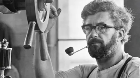 Black and white image of Stanley Graham, with curly hair and a beard, wearing gasses and with a pipe in his mouth, holding a wheel in the mill engine house in 1976