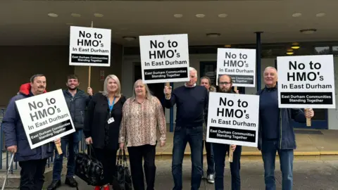 A group of residents are stood carrying placards saying 'No to HMO's in East Durham'. In smaller writing at the bottom it reads 'East Durham Communities Standing Together'