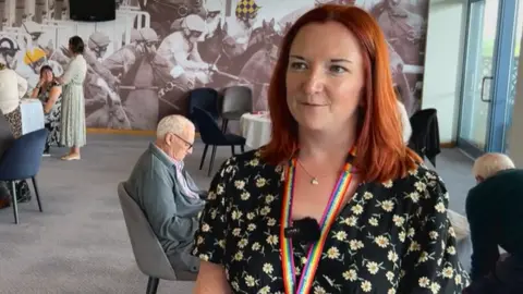 A woman with red hair, wearing a black floral dress and rainbow lanyard stands in front of the camera