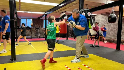 Un niño con un chaleco verde, pantalones cortos negros y botas rojas golpea una almohadilla manual sostenida por un hombre barbudo con una camiseta azul y pantalones crema, con otros boxeadores jóvenes en el fondo
