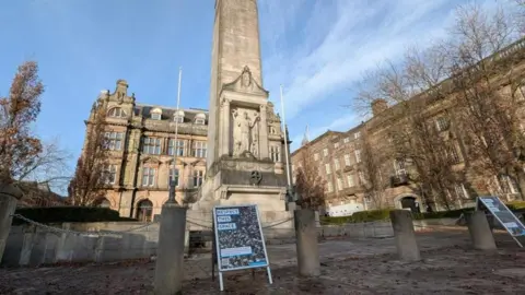 Two A-boards saying 'Respect this space' are placed in front of the stone columns and chains that surround the Cenotaph in Preston. 