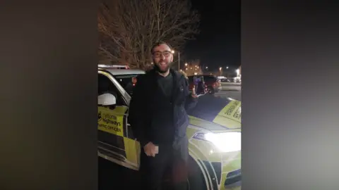 A man smiles at the camera as he holds up a mobile phone. He is standing in front of a National Highways' car that is parked on the road. The photo is taken at night. 