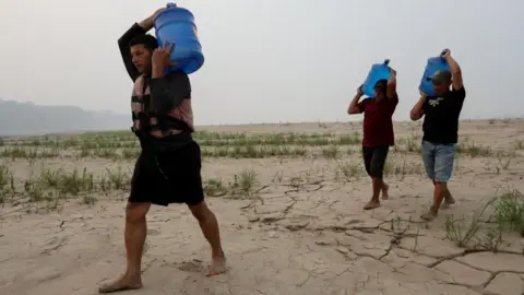 Three men carry drinking water above their heads in large blue plastic tanks along a dry sandbank of the Madeira River in northern Brazil