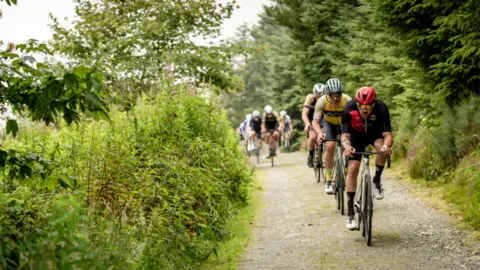 On a green lane, a line of cyclists ride gravel bikes