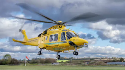 A yellow helicopter is taking off from a field in front of a cloudy sky.
