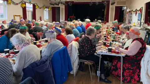 Becki Bowden / BBC A wide shot of the elderly people enjoying the Christmas lunch. They are seated at rows of long tables. 