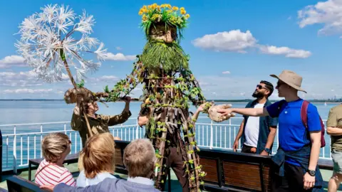 The scene is set outside under a bright blue sky with scattered, wispy white clouds. The lighting suggests daytime with strong natural sunlight. A crowd has gathered, watching a performance happening on a small raised structure. The performer is dressed in plants and flowerbeds.