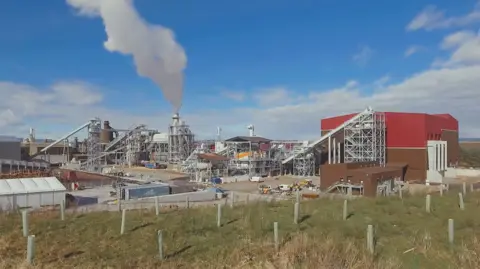 A large industrial site near Inverness with smoke rising from a chimney. There are several different buildings including a large bright red warehouse.