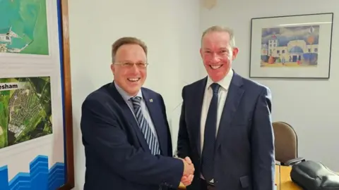 Michael Keohan/BBC Charles Woodgate (left) shakes hands with Conservative opposition leader Trevor Bartlett (right) after defecting to the Conservatives from Labour