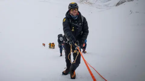 Tim Howell is climbing up a mountain holding on to rope. He is wearing weather protective gear. There is a team of people behind him.