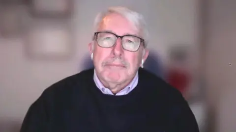 Chris Blythe in a black jumper and purple top. He is wearing glasses and airpods. Behind him his background is blurred. 