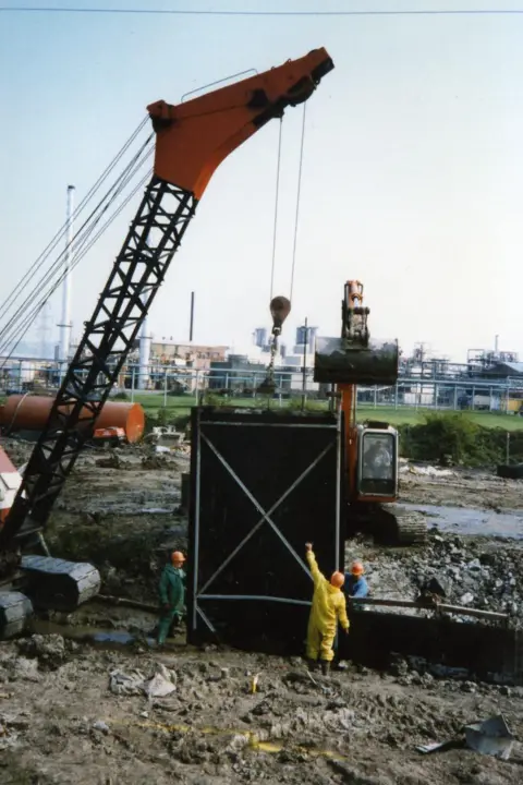 Stephen Constance A work site where a crane is lowering something in a black container into a hole. There are workers wearing overalls and orange hats stood around the site.