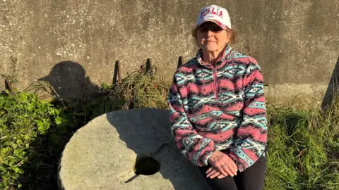 Marie Howden sits on a millstone with a concrete wall behind her. She is wearing a colourful jumper, a baseball cap and glasses.