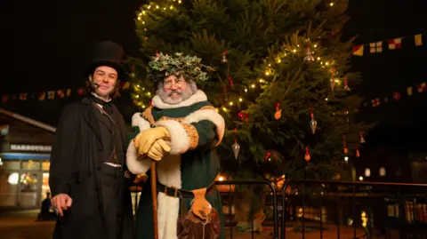 SS Great Britain Two actors dressed up as a green Father Christmas and what looks like Isambard Kingdom Brunel in a top hat and with a pipe. Both are standing in front of a big Christmas tree, smiling at the camera. 