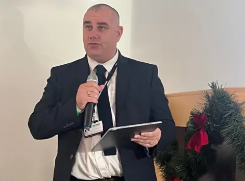 Great Yarmouth Borough Council A man holding a microphone while wearing a suit speaks at a conference in a white room while holding a tablet.