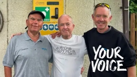 Neil Berrigan Jim Berrigan has his arms around  Steven and Graeme as he stands in front of the defibrillator box which is fastened to the wall of a building. 
