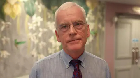 A man, with short white hair, wearing glasses, a dark red tie and a blue shirt. 