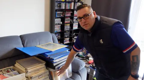 Stefan Bennett has some of the donated items at his house, and is posing in front of boxes full of old programmes in his living room, wearing a Gillingham gilet