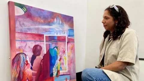 A woman with white sunglasses on her head kneeling next to a multi-coloured painting 