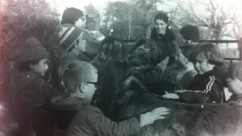 Farms for City Children The picture shows a group of children gathered around a large, shaggy‑coated animal, most likely a donkey or pony, in an outdoor farm setting. The children are reaching out to stroke the animal, some leaning in close, while others stand nearby watching. The scene appears to be an old black‑and‑white photograph.