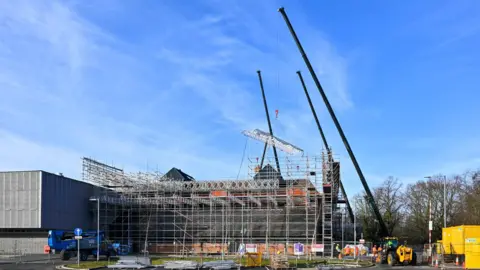 Stuart Walker The Sands Centre which is a grey building covered with scaffolding. Three black cranes are beside it.