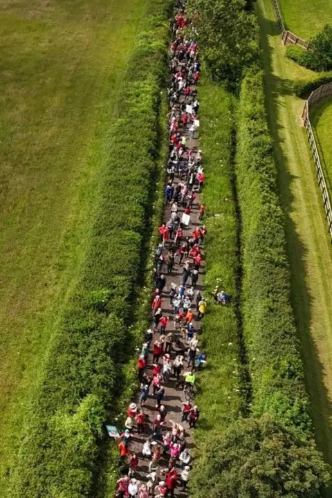 Stop Lime Down Hundreds of people are walking along a road in the middle of the countryside holding banners. The picture has been taken from a drone.