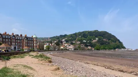 BBC A beach with a promenade and buildings to the left. North Hill is to the right of the image. There are dark and pale green trees on a hill. The picture was taken on a sunny day.