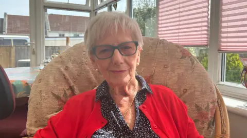Pauline Heyworth pictured with short grey hair and green glasses. She is wearing a red cardigan with a navy blouse on underneath. Pauline is wearing pearl earrings and is smiling with closed lips.