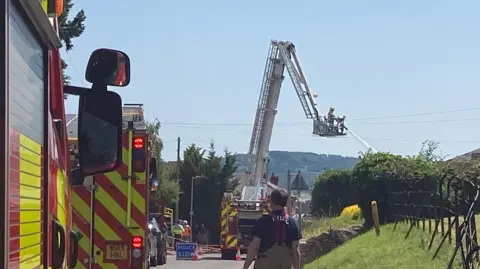 Fire crews shoot water out of a hose as they stand on an aerial device, much like a cherry picker, with fire engines on the road below. There are police signs and there are emergency service workers walking down the road.