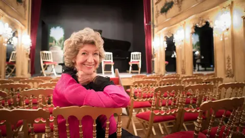 Dame Felicity Lott with short curly hair dressed in black and pink leans on the back of a chair and smiles to a camera while sitting in a theatre space.
