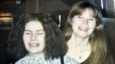 Diane Barnes An old photo of two young women on a night out. They are grinning at the camera and standing beside each other. One has dark hair and the other has brown hair. They are standing in a darkly lit indoor venue. 