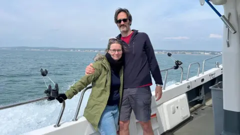 Joshua Askew/BBC A man and woman stand arm in arm on a boat.