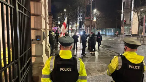 The back of two police officers looking in the direction of a small group of protestors all dressed in dark clothing standing on a footpath. One is holding an England flag