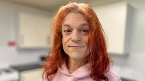 Martin Giles/BBC Emma is looking at the camera she has long red hair and is wearing a pink hoodie.