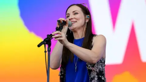 Julie Fowlis has long dark hair and sunglasses perched on top of her head. She is smiling as she sings into a microphone fitted to a microphone stand.