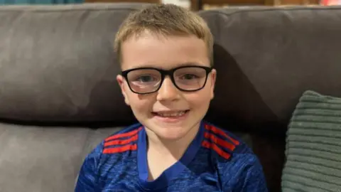 BBC Mabon in a blue football short and glasses sits on a settee smiling at the camera. It is a head and shoulders shot.